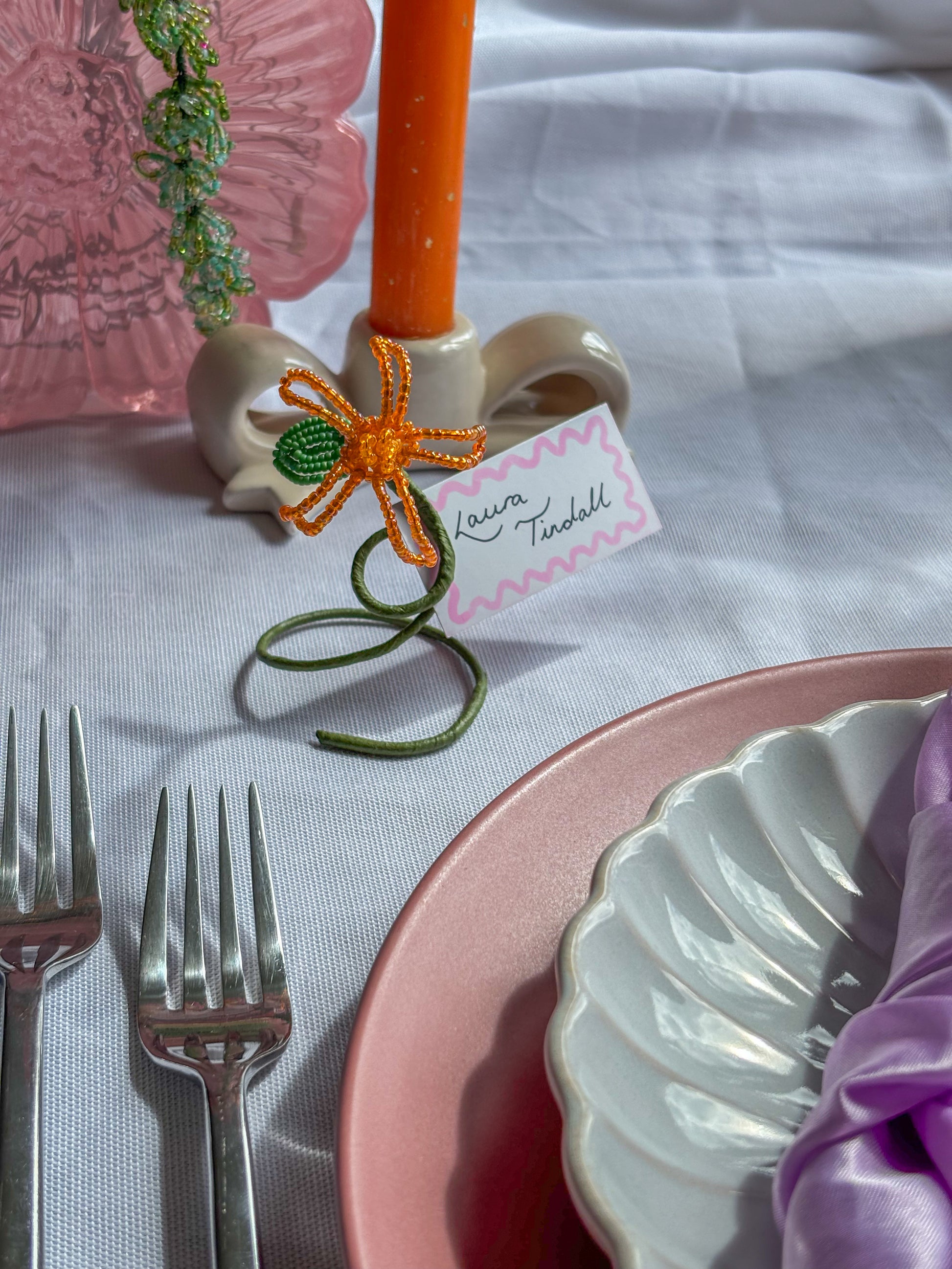 Table setting with pink plates, silverware, and a decorative name card holder with an orange flower.